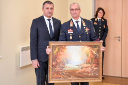 A képen az országos katasztrófavédelmi főigazgató és Vass Gyula az ajándékba kapott festménnyel, a háttérben a személyzeti főosztály vezetője.